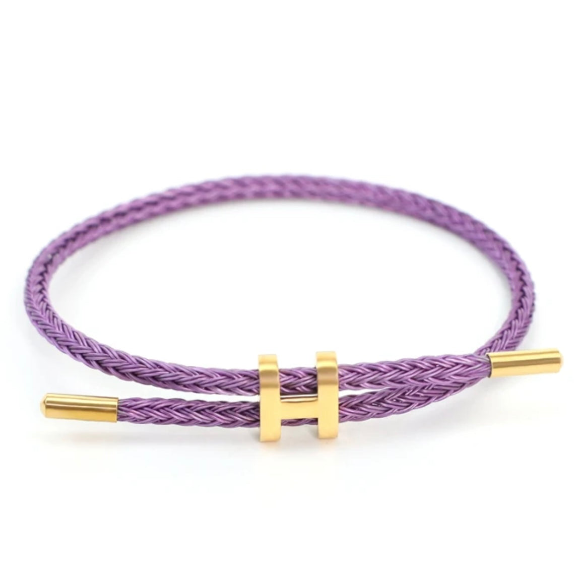 A studio photo of a minimalist purple bracelet. The bracelet is made of a braided purple rope and features a sleek gold-toned metal clasp in the shape of a capital 'H' that can be adjusted for size. The bracelet is displayed on a clean white background.
