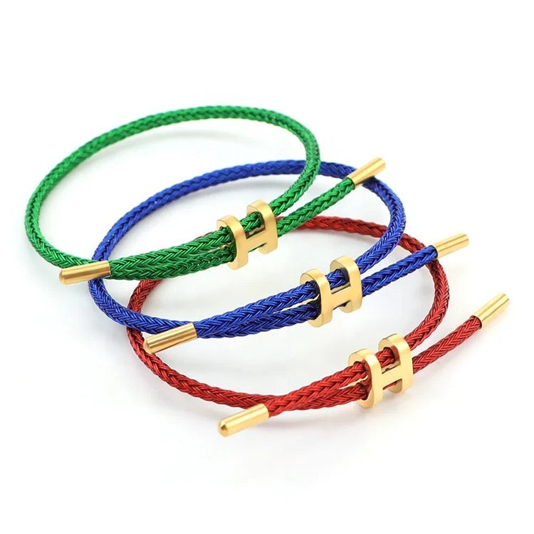 A product photo of three adjustable bracelets stacked together. The bracelets are made of braided rope in primary colors: vibrant green, royal blue, and red. Each bracelet features a polished gold-toned metal clasp in the shape of a capital 'H'. The set is displayed on a white background.