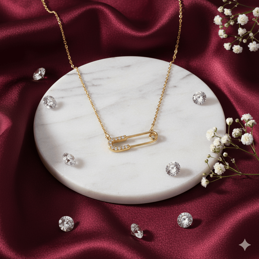Gold necklace featuring a safety pin pendant encrusted with small diamonds, displayed on a white marble base, surrounded by loose gemstones and white flowers, set against an elegant maroon silk background.