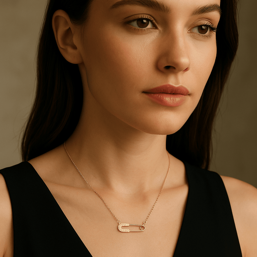 Gold necklace featuring a safety pin pendant encrusted with small diamonds, worn around the neck of a woman with dark hair and a black top, highlighting the jewelry's elegance.